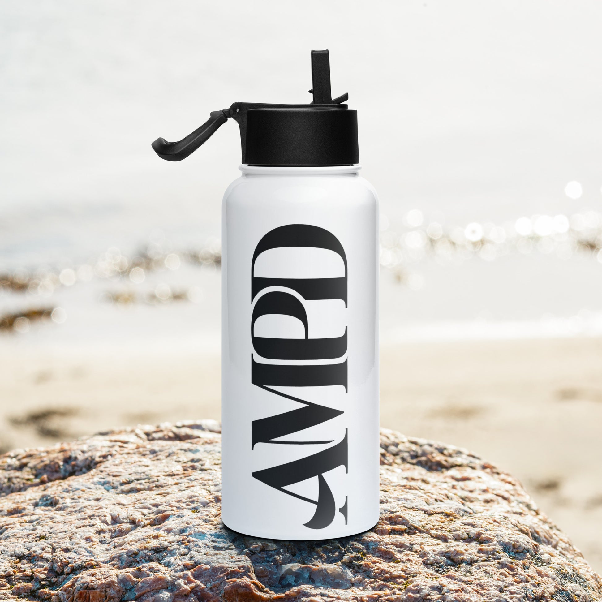 AMPD Stainless steel water bottle with a straw lid on a rock by the beach