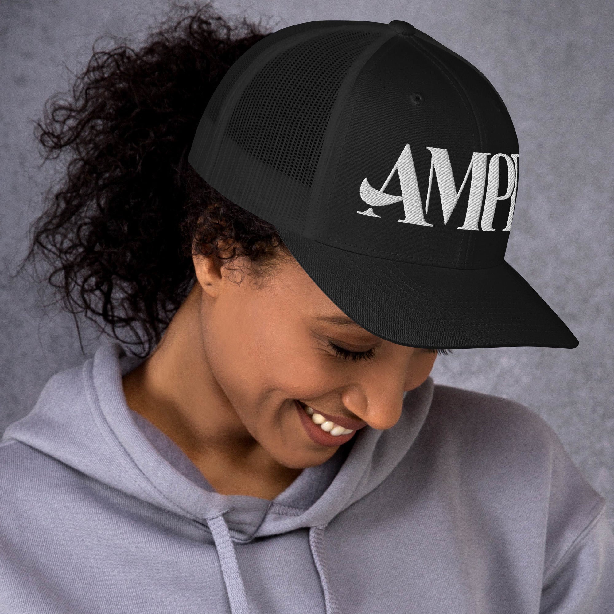 Woman wearing an Amp'd Up Logo Trucker Cap in black, styled with a casual hoodie